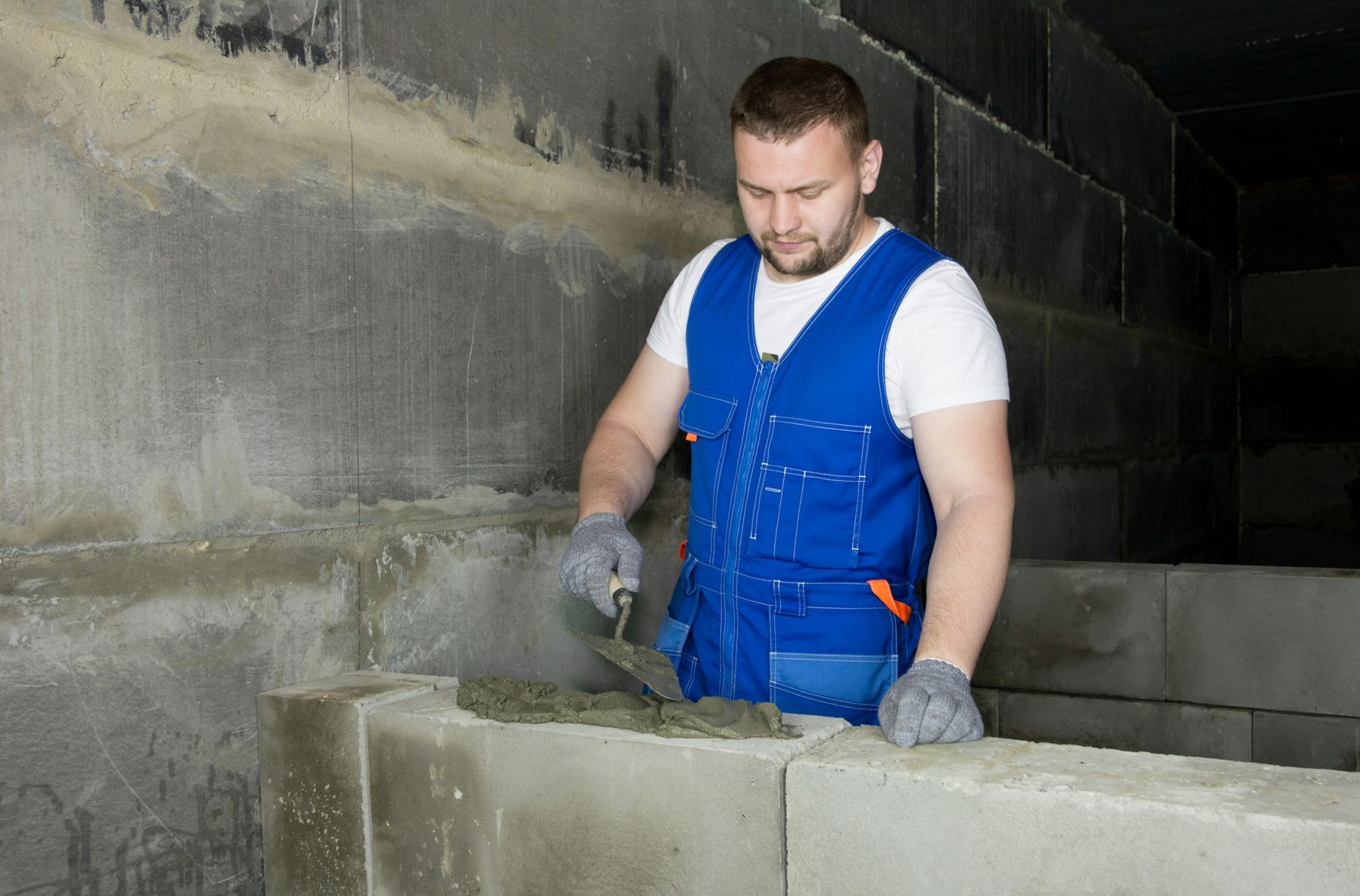 A builder in blue overalls laying cement on concrete blocks
